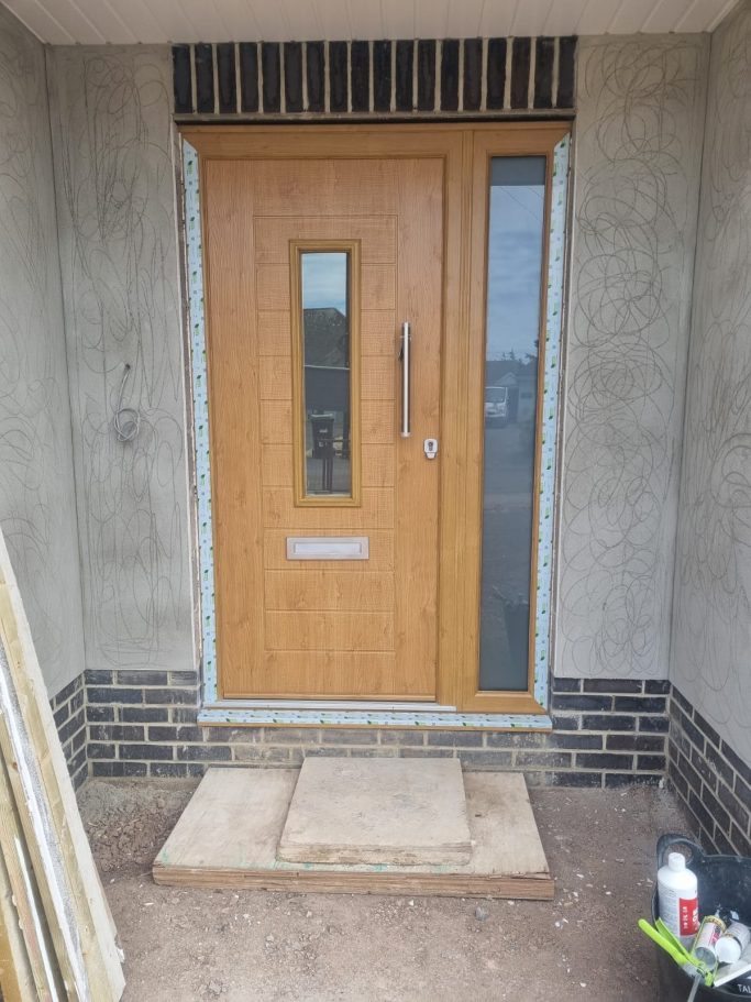 A modern wooden front door with a glass panel, framed by unfinished walls and bricks.